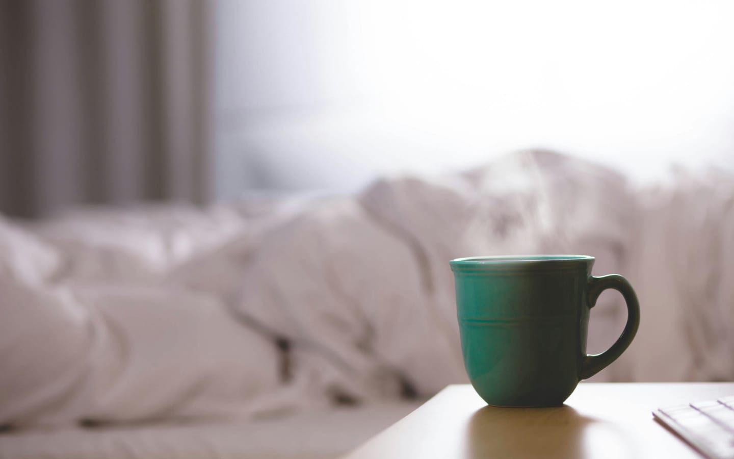 Cozy morning with a steaming green coffee mug on a bedside table, a blurred bed in the background, and a warm, relaxing vibe